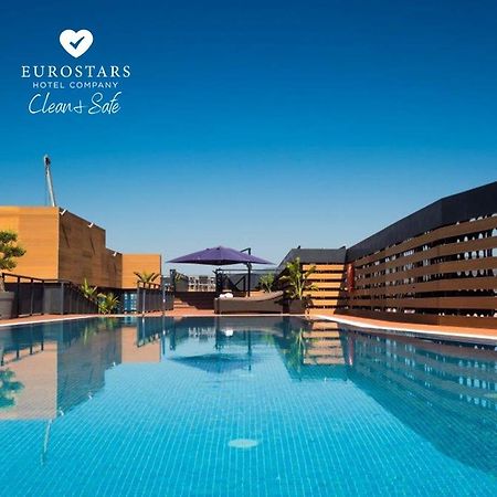 Eurostars Palace Cordoba Ngoại thất bức ảnh The photo shows an outdoor swimming pool with clear, blue water, reflecting the blue sky above. The pool area is surrounded by modern architectural features, including wooden slats and potted plants. There is also a purple umbrella providing shade. T
