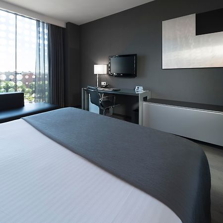 Eurostars Palace Cordoba Ngoại thất bức ảnh The photo shows a modern hotel room featuring a sleek design. In the foreground, there is a bed with a white coverlet and gray accents. To the side, a dark sofa is visible. The room has a large window with sheer curtains, allowing natural light to en