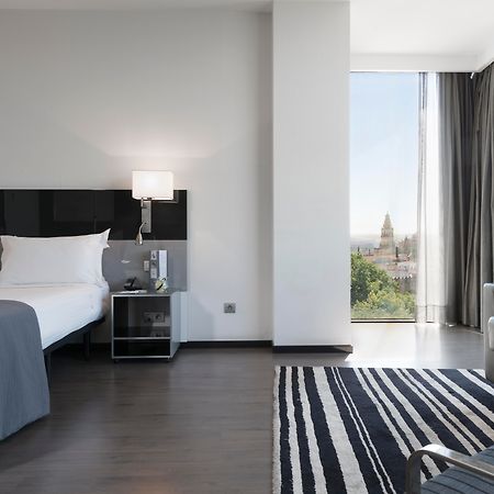 Eurostars Palace Cordoba Ngoại thất bức ảnh The photo shows a modern hotel room featuring a large bed with white linens and dark cushions, paired with a sleek nightstand. There is a contemporary chair and a small table nearby, along with a striped area rug on the floor. A large window allows n
