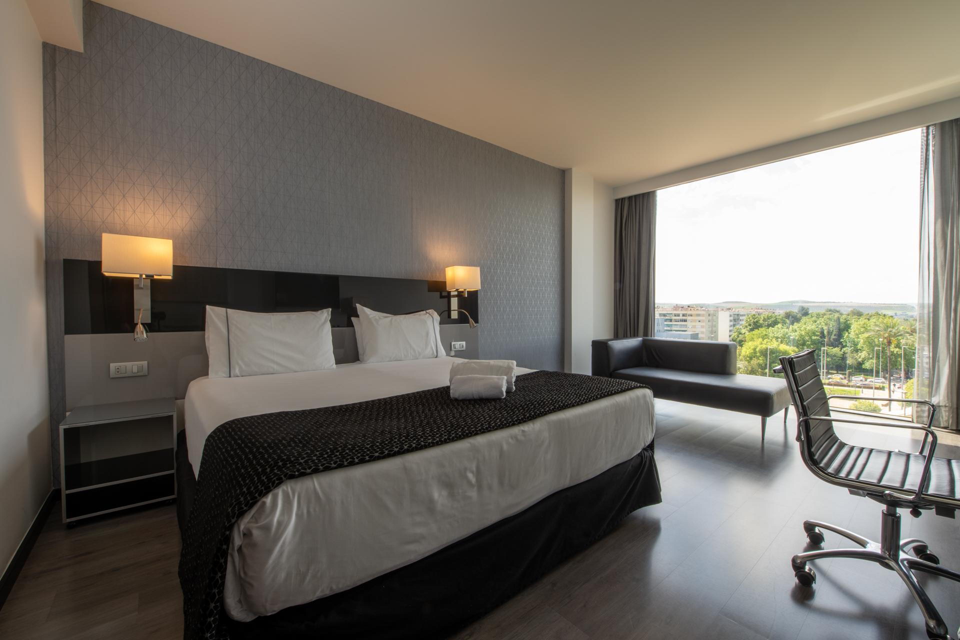 Eurostars Palace Cordoba Ngoại thất bức ảnh The photo shows a modern hotel room featuring a large bed with white linens and a dark patterned throw. On either side of the bed, there are stylish wall-mounted lamps. A contemporary desk chair is positioned at a small desk, and there’s a sofa near 