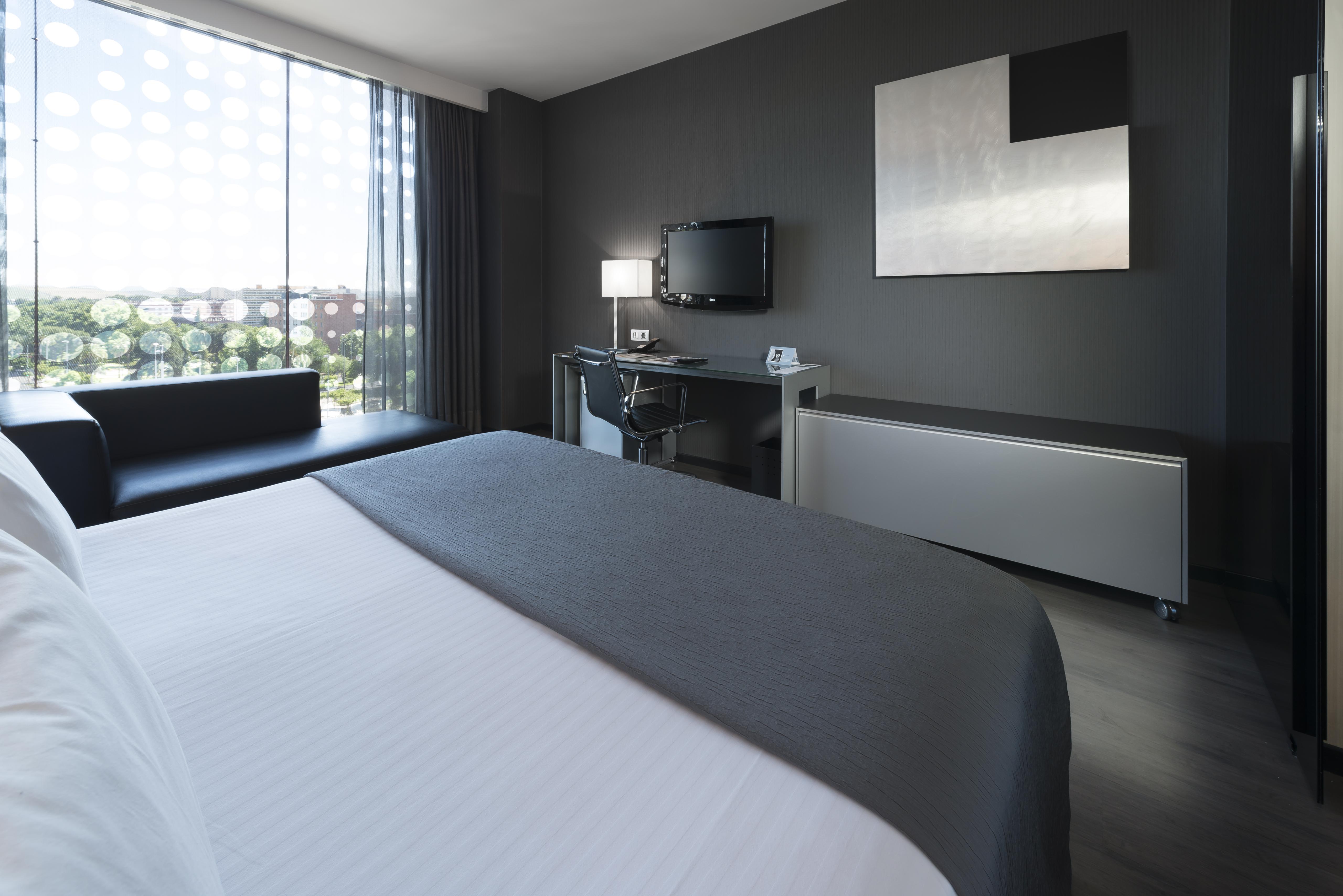 Eurostars Palace Cordoba Ngoại thất bức ảnh The photo shows a modern hotel room featuring a sleek design. In the foreground, there is a bed with a white coverlet and gray accents. To the side, a dark sofa is visible. The room has a large window with sheer curtains, allowing natural light to en