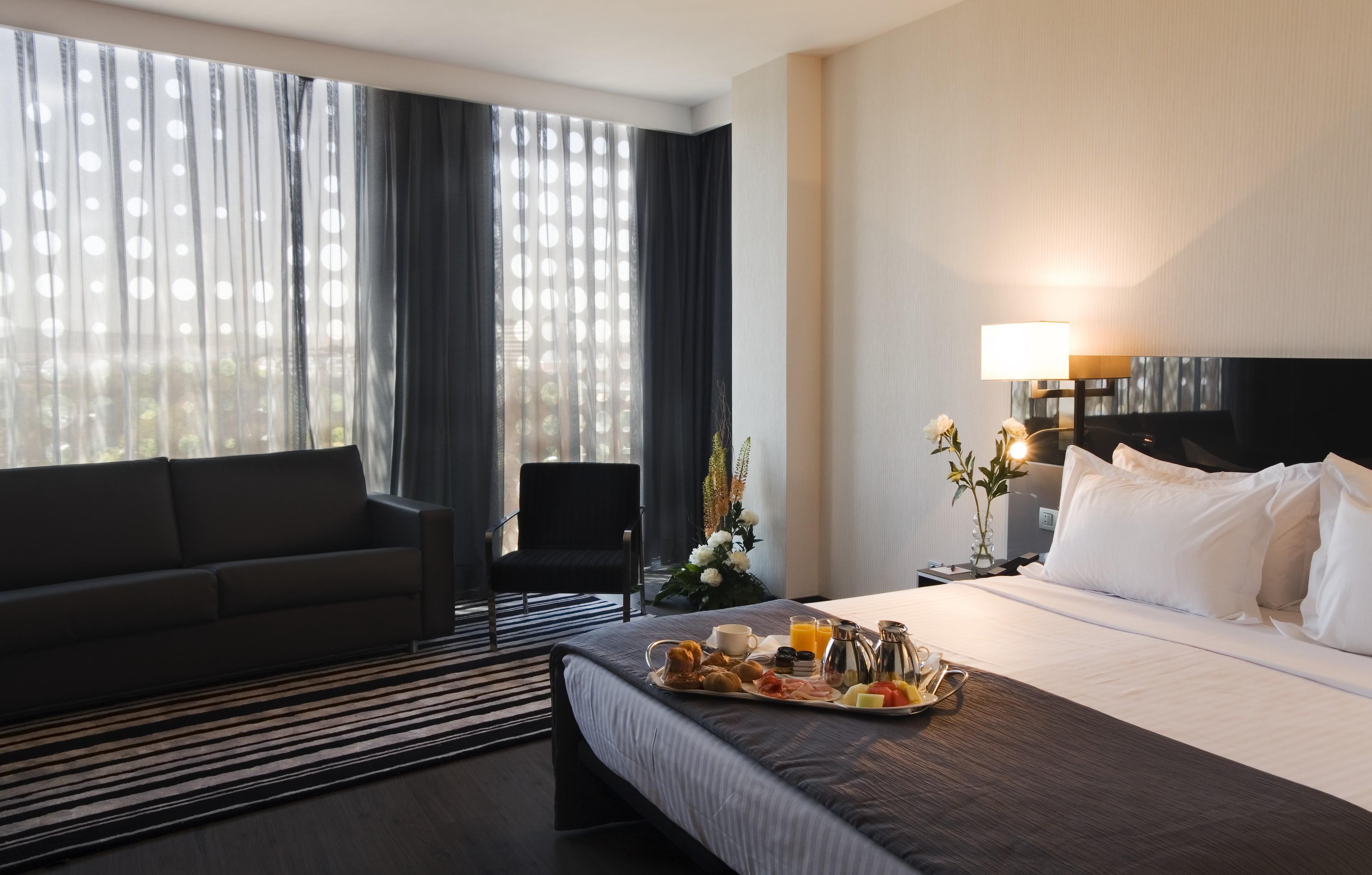 Eurostars Palace Cordoba Ngoại thất bức ảnh The photo shows a modern hotel room featuring a neatly made bed with white linens and a dark grey blanket. There is a tray on the bed with a breakfast spread, including pastries, fruit, and beverages. To the left, there is a comfortable-looking couch