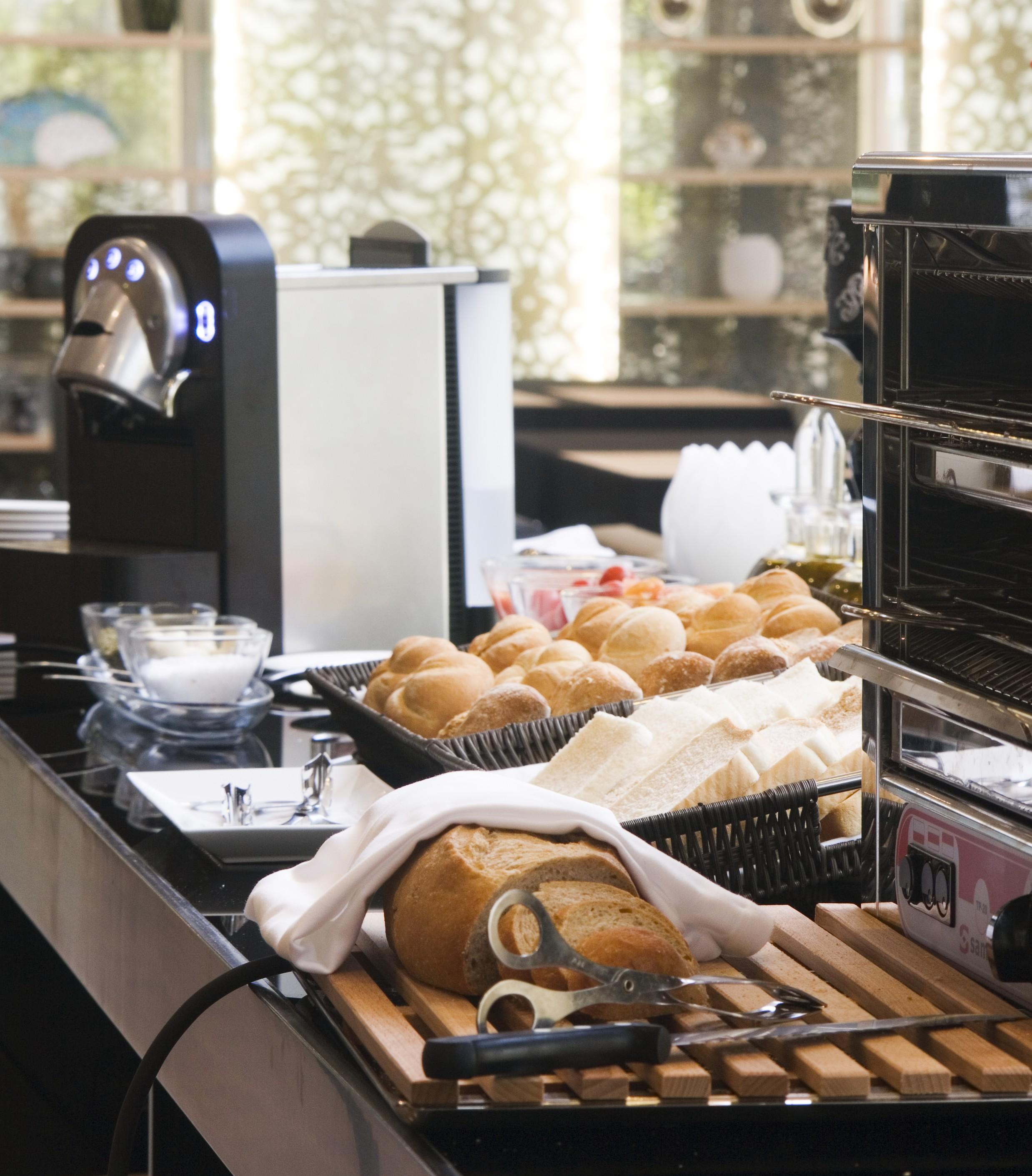 Eurostars Palace Cordoba Ngoại thất bức ảnh The photo shows a dining setup featuring various food items. On the left, there is a coffee machine, and in the foreground, there is a basket of freshly baked bread rolls. Next to the rolls, there are slices of bread arranged neatly. In the backgroun