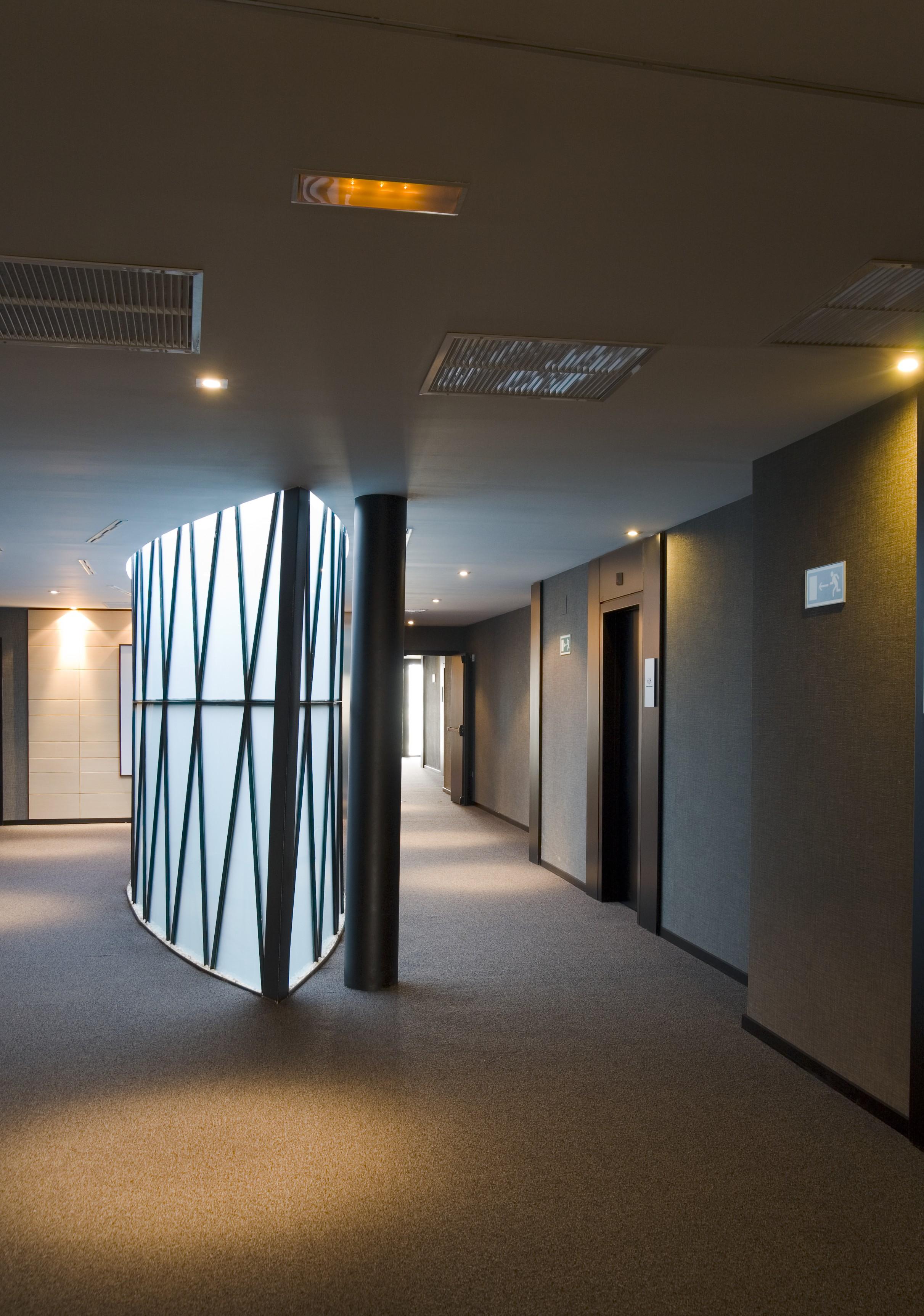 Eurostars Palace Cordoba Ngoại thất bức ảnh The photo shows a modern indoor corridor featuring a smooth, curved wall with a geometric pattern. The corridor is well-lit with recessed ceiling lights, and the floor is carpeted. There are doors leading to elevators on the right side, and the walls