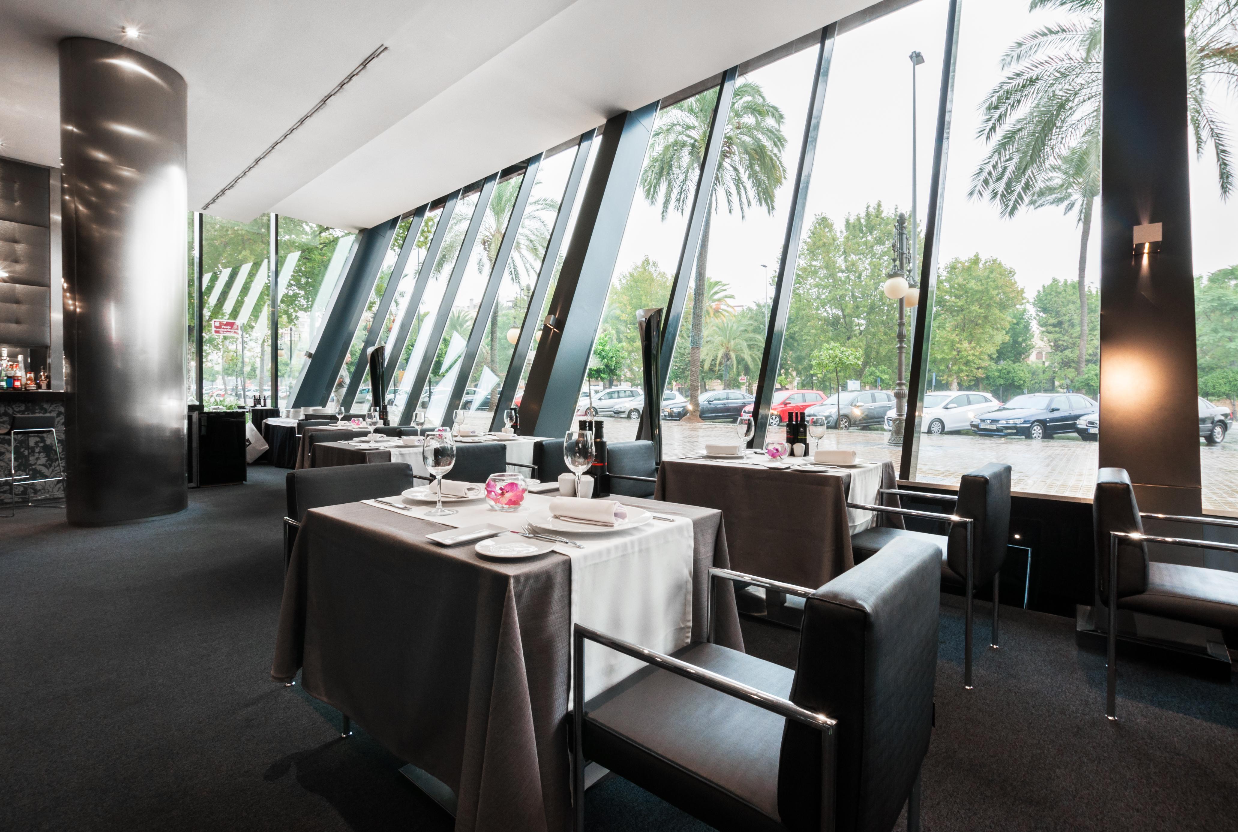 Eurostars Palace Cordoba Ngoại thất bức ảnh The photo shows the interior of a modern restaurant featuring large glass windows that provide an expansive view of the outdoors, including palm trees. The dining area is elegantly set up with tables covered in gray tablecloths, decorated with simple