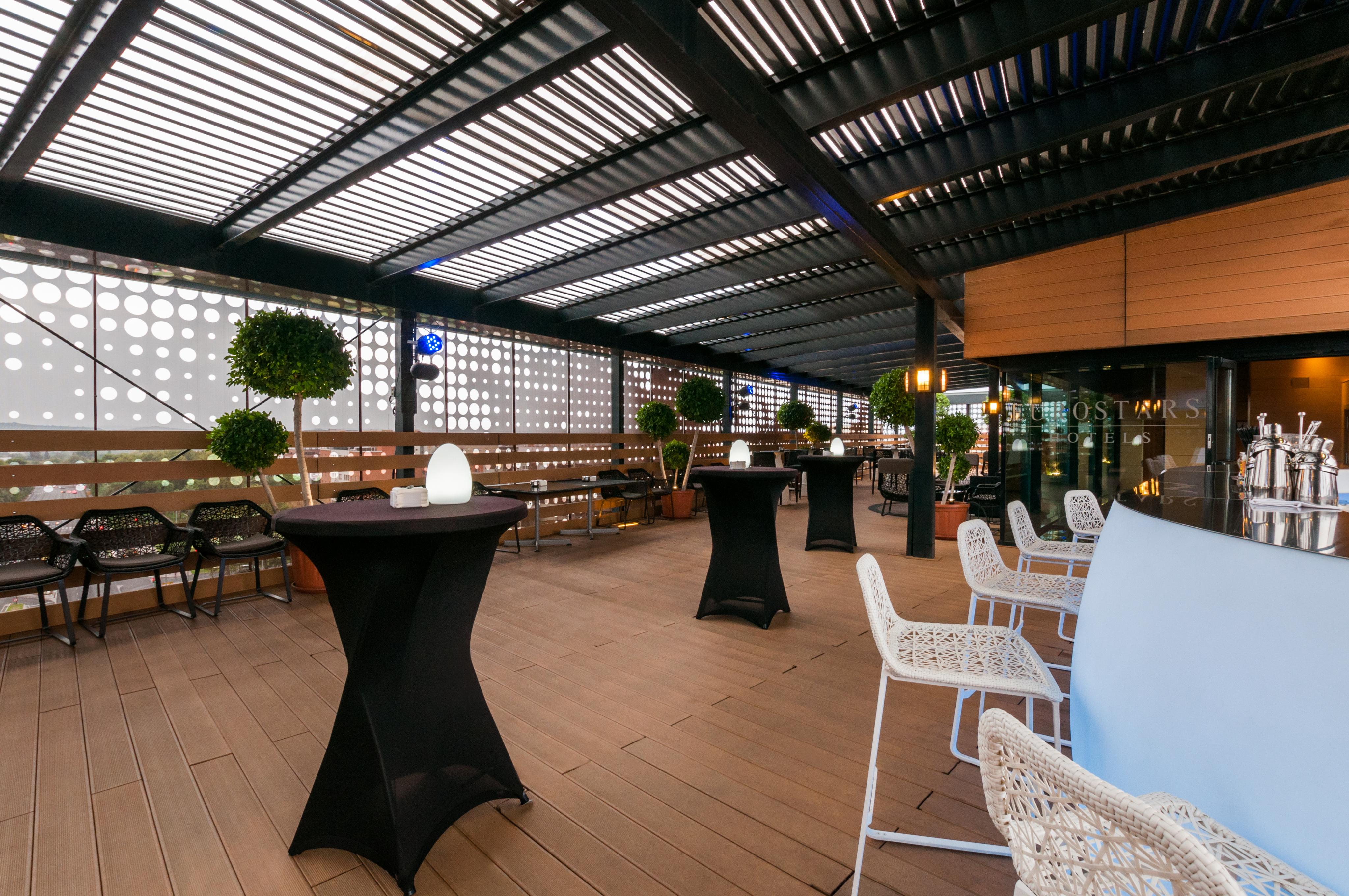 Eurostars Palace Cordoba Ngoại thất bức ảnh The photo shows a stylish outdoor seating area, likely part of a restaurant or lounge. It features a wooden floor and an overhead structure with horizontal slats, allowing for both shade and light. There are several tall, black cocktail tables and wh