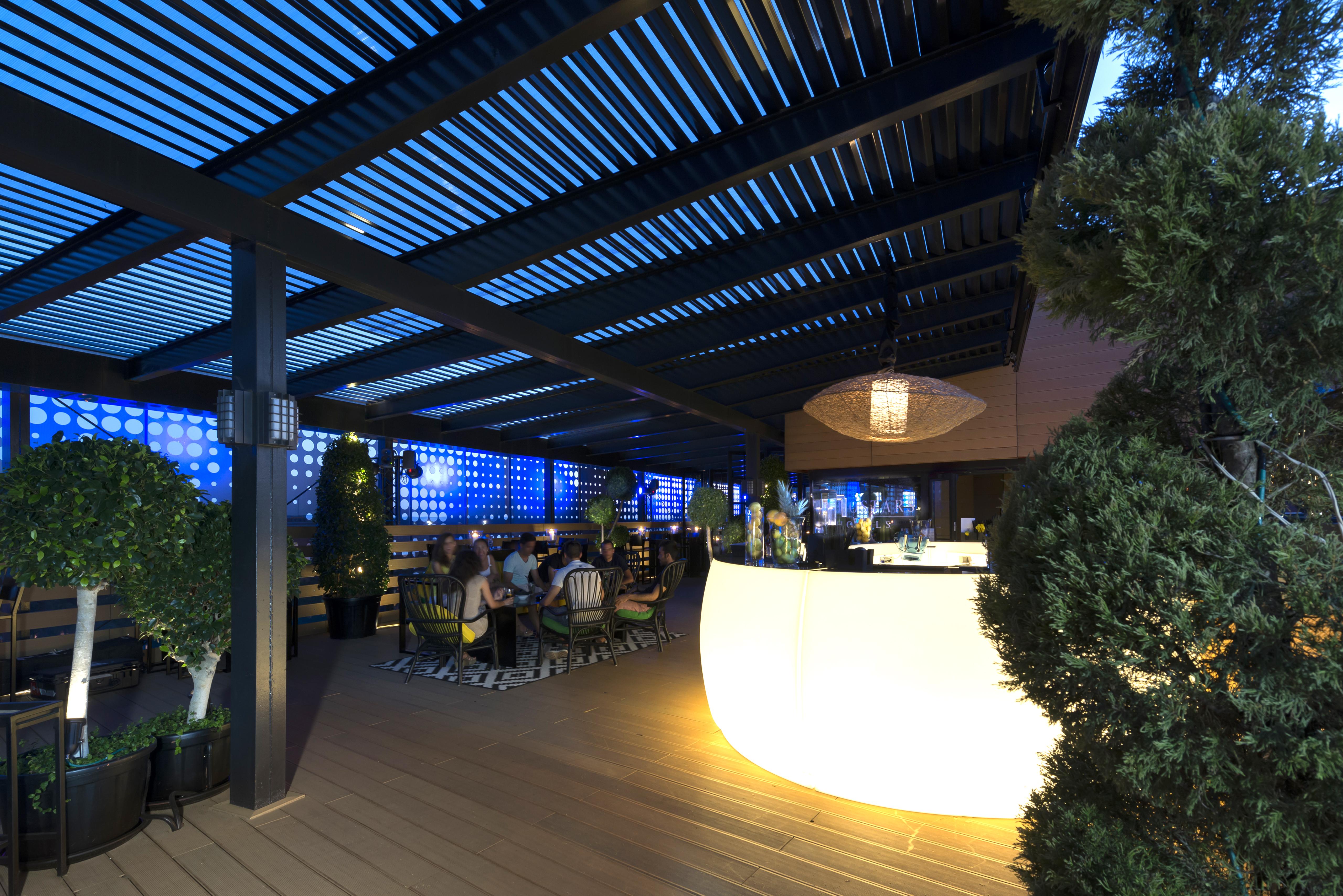 Eurostars Palace Cordoba Ngoại thất bức ảnh The image shows a modern outdoor bar or restaurant area. The space features a sleek, illuminated bar that stands out with its curved shape. Patrons can be seen seated around tables, enjoying the ambiance. The overhead structure consists of slatted pa