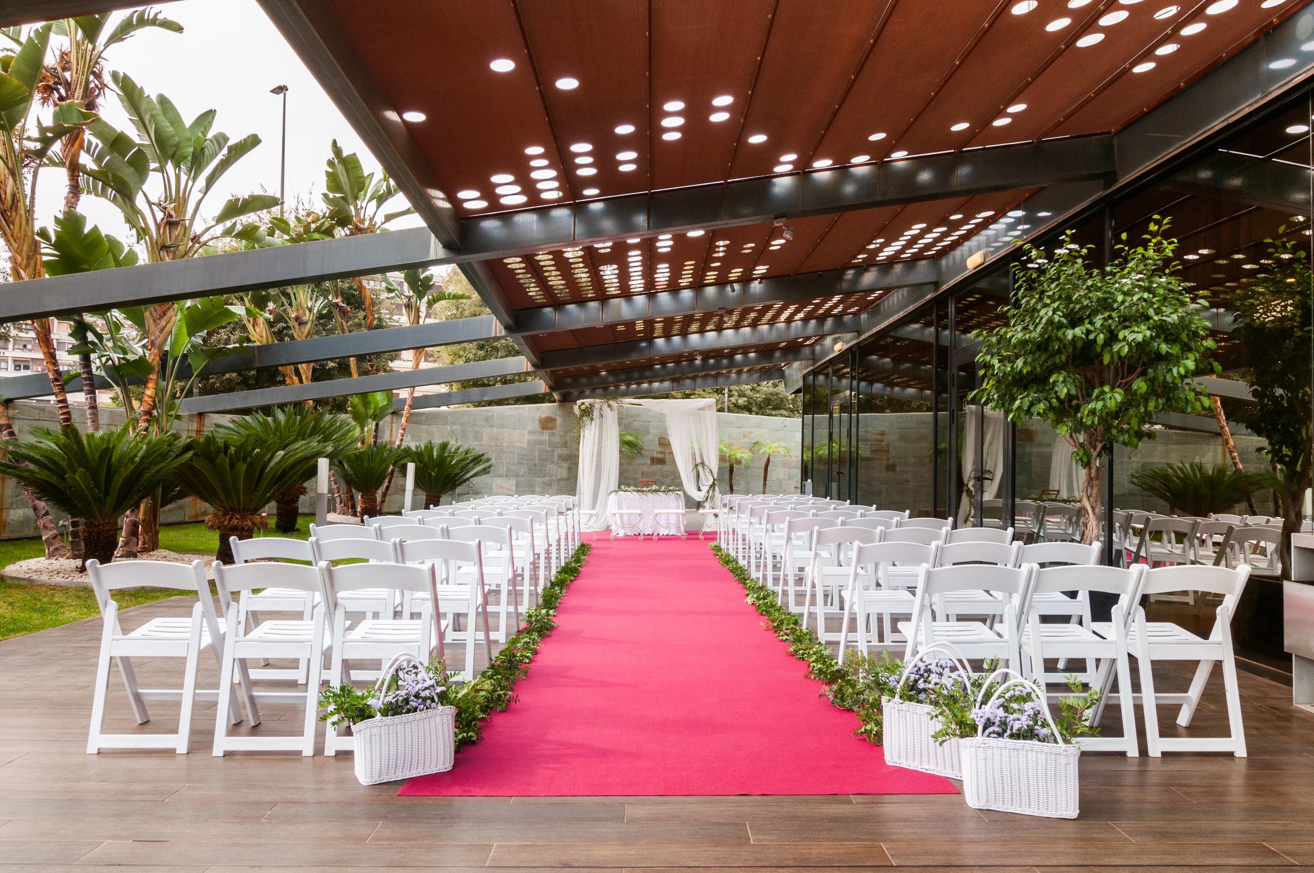 Eurostars Palace Cordoba Ngoại thất bức ảnh The photo shows a wedding setup in an outdoor venue. There are white chairs arranged in rows on either side of a vibrant pink carpet leading to a floral arch or backdrop at the front. The area is partially shaded by a modern structure with a metal fr