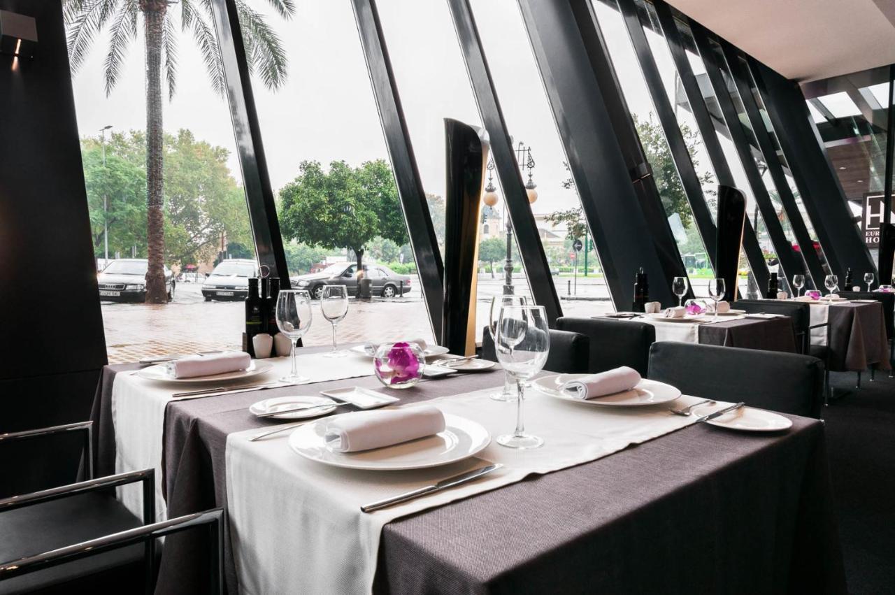 Eurostars Palace Cordoba Ngoại thất bức ảnh The photo shows an elegant restaurant interior with large windows featuring modern angular designs. There are two dining tables set up, each covered with white and dark tablecloths. The tables are elegantly arranged with plates, cutlery, and wine gla