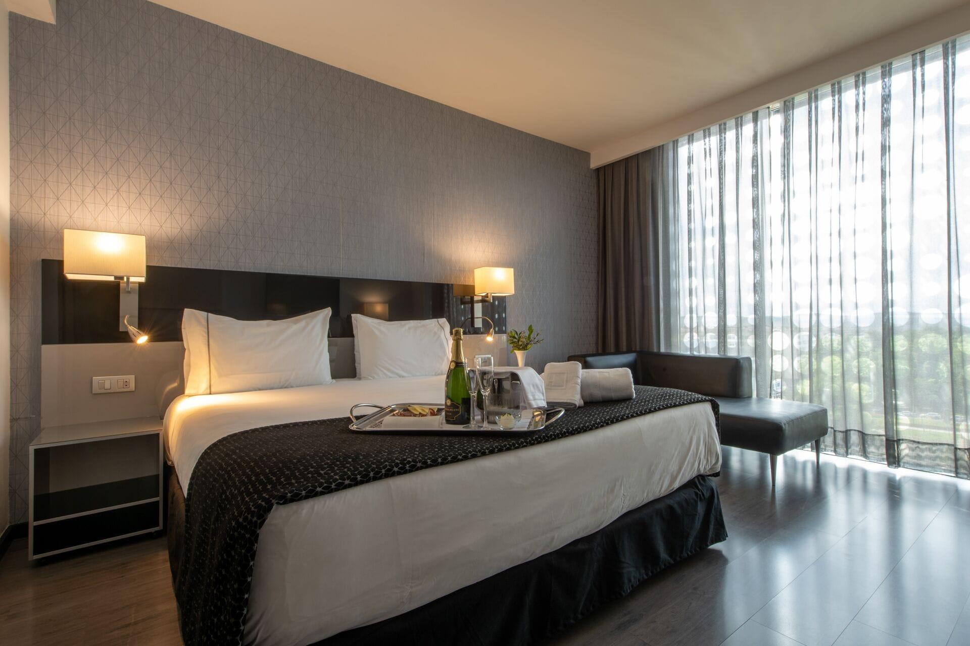 Eurostars Palace Cordoba Ngoại thất bức ảnh The photo shows a modern hotel room featuring a large bed with crisp white linens and dark throw blankets. On either side of the bed, there are bedside lamps providing soft lighting. The room has a sleek design with a neutral color palette, including