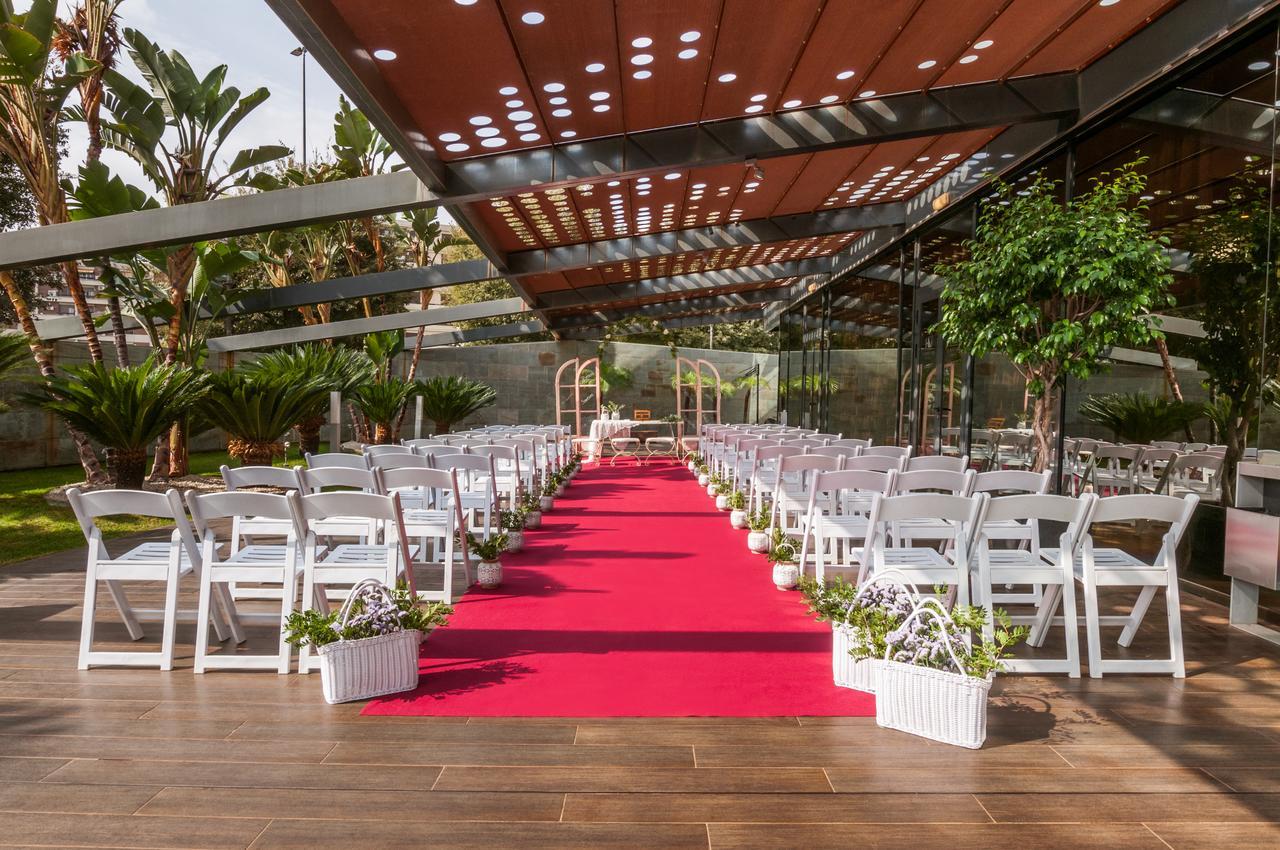Eurostars Palace Cordoba Ngoại thất bức ảnh The image shows an outdoor wedding setup. There are white chairs arranged neatly on either side of a vibrant red carpet that leads to a framed archway. The area is covered by a glass and metal structure, allowing plenty of natural light, while tropic