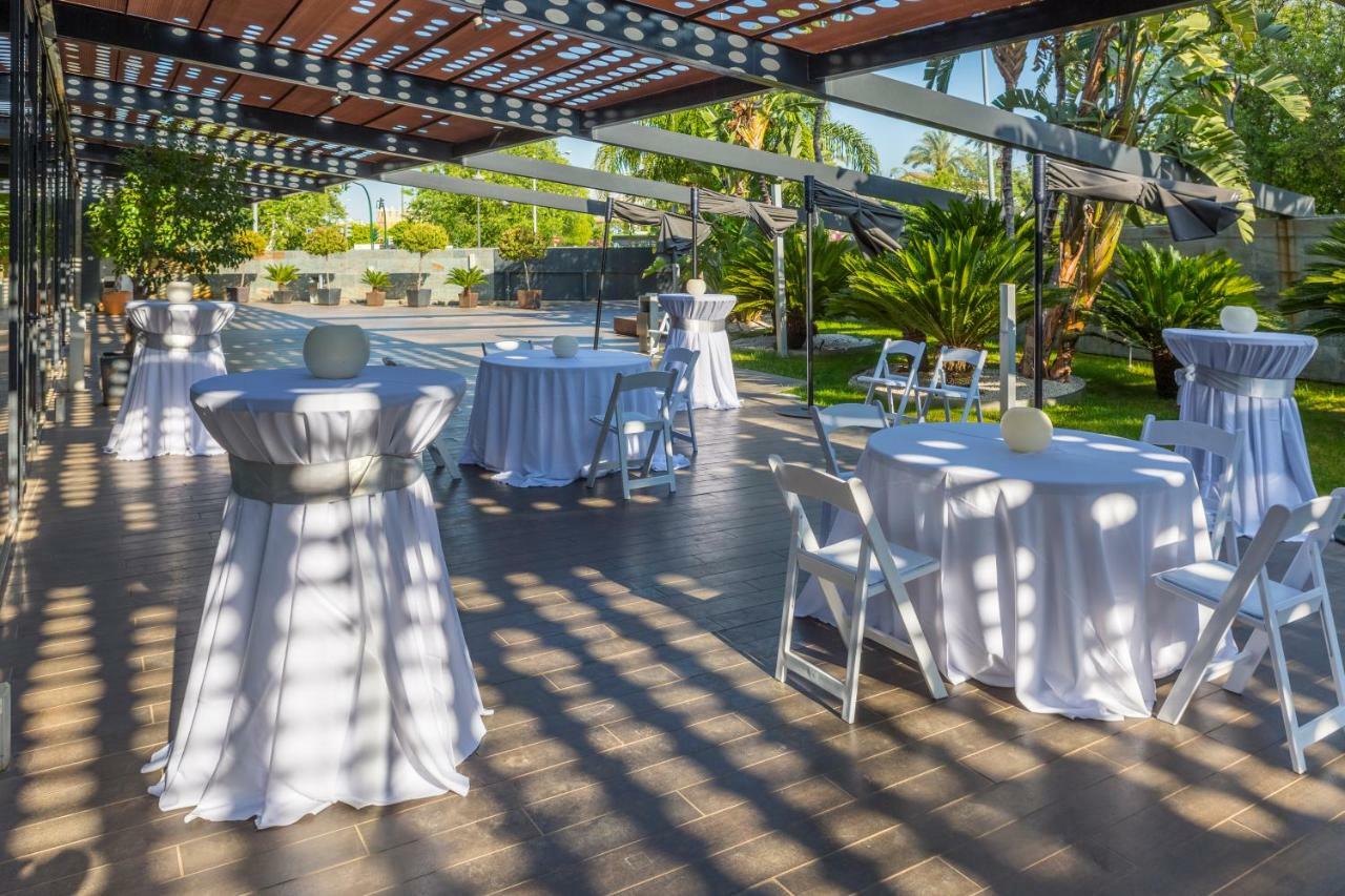 Eurostars Palace Cordoba Ngoại thất bức ảnh The photo shows an outdoor event setup. There are several round tables covered with white tablecloths, some adorned with decorative elements like small centerpieces. Chairs are arranged around the tables, and the area is surrounded by greenery, inclu